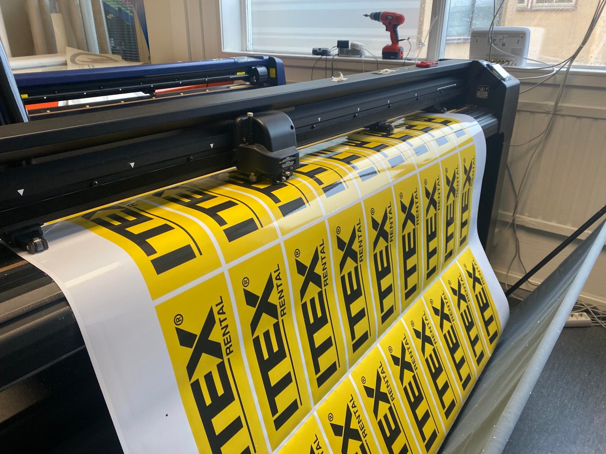 Storformatsskrivare som producerar gula dekaler med svart text och logotyp för HEX Rental. En lång rulle självhäftande vinyl matas genom maskinen och visar upprepade, jämnt placerade dekaler i rutmönster. Maskinen står i en verkstadsmiljö med fönster, verktyg och kablar synliga i bakgrunden, vilket signalerar pågående skylt- och dekalproduktion.