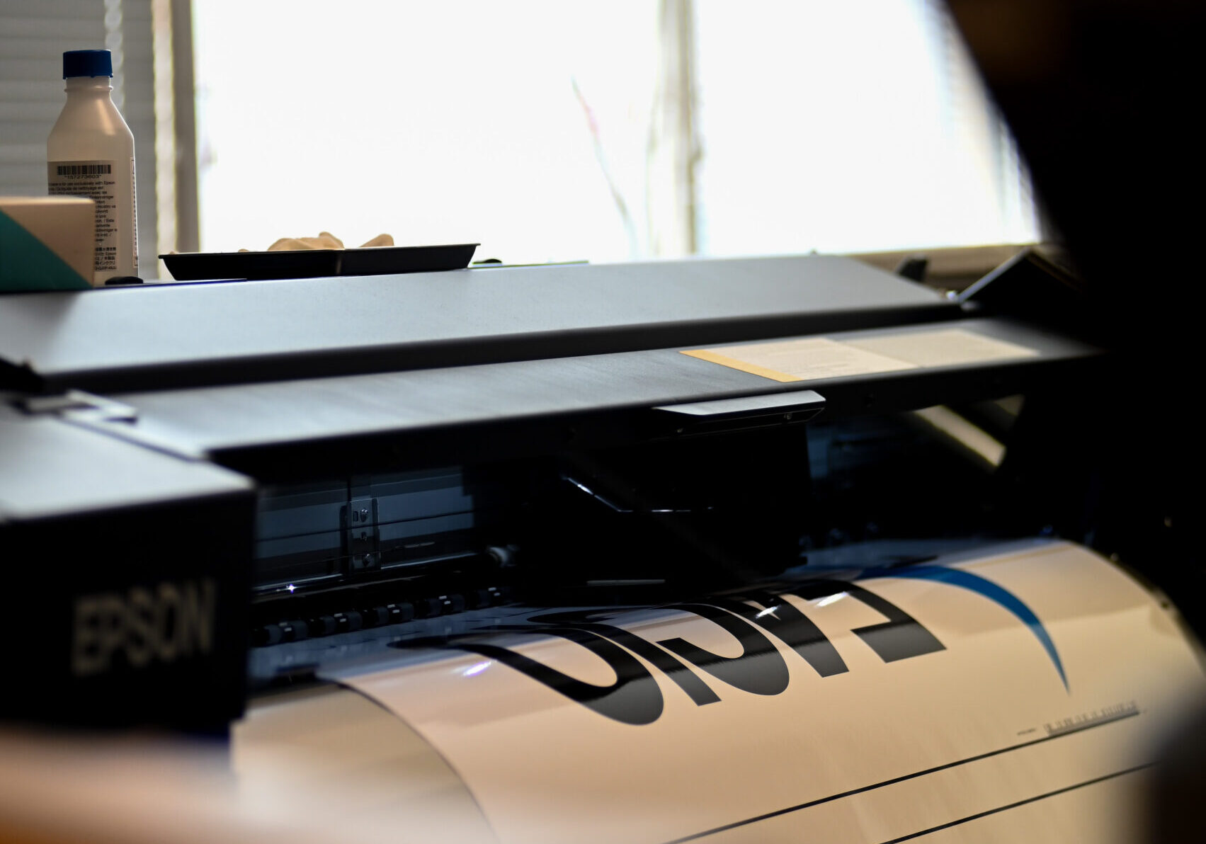 En stor bläckstråleskrivare i en ljus arbetsmiljö håller på att skriva ut en affisch eller skylt på rulle. Pappret matas ut från maskinen med tydlig svart typografi och grafiska former. I bakgrunden syns ett fönster med persienner som släpper in dagsljus, samt en flaska och små föremål placerade ovanpå skrivaren, vilket ger intryck av pågående produktion i en verkstad eller ateljé.